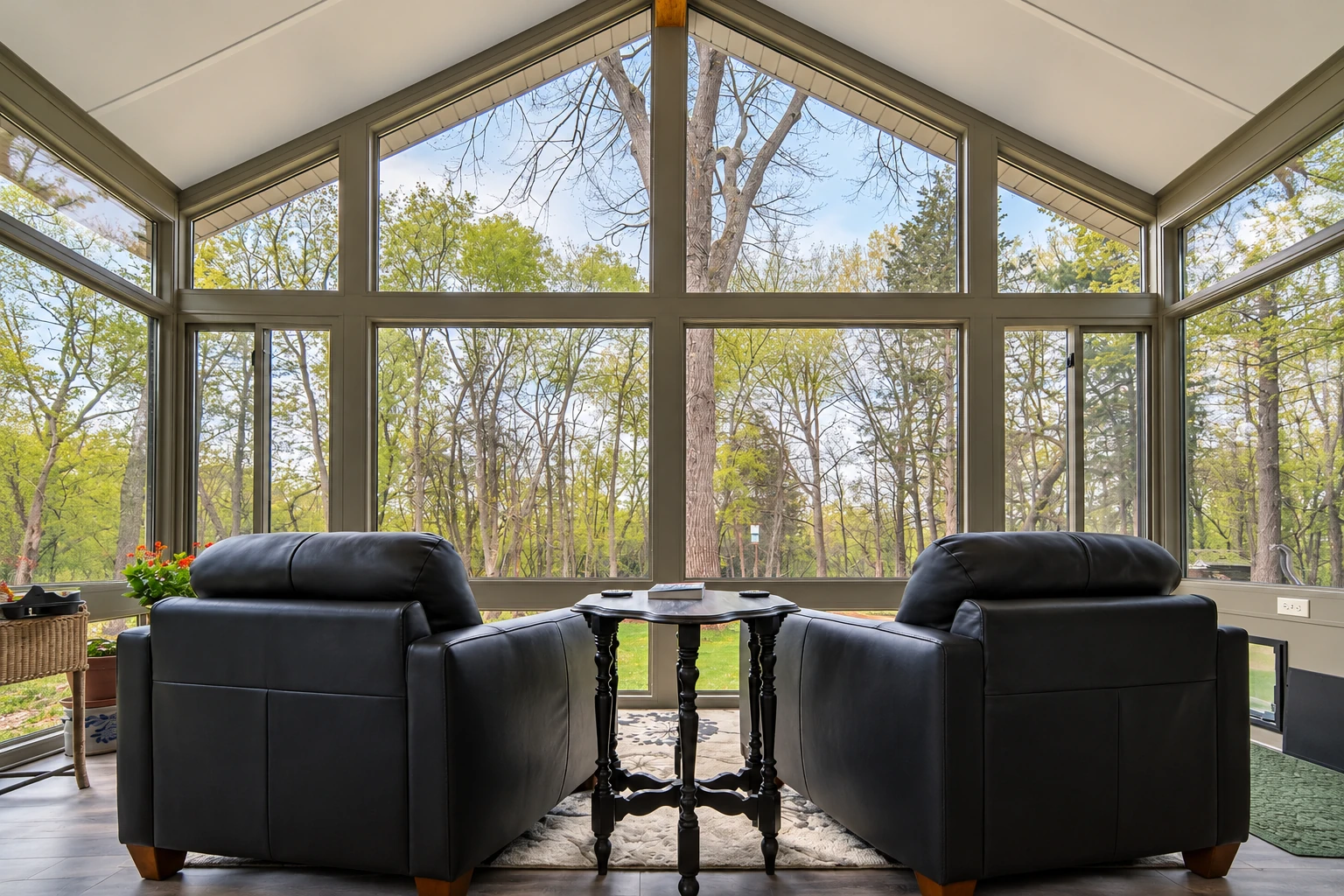 A four-season sunroom has tall, olive-framed windows and two black leather armchairs facing a small table, with trees outside.