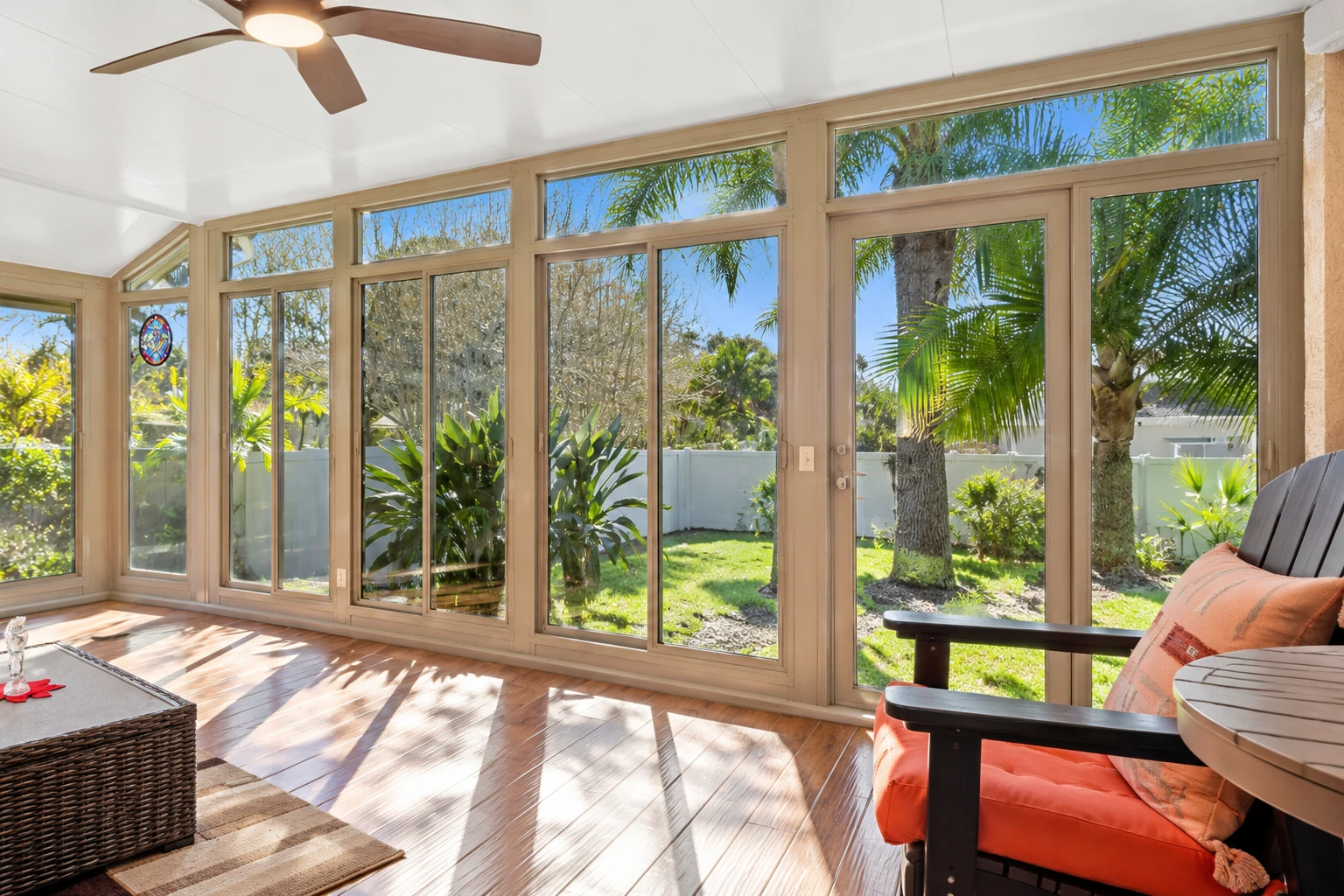 Sunroom addition with tan-framed floor-to-ceiling windows, a ceiling fan, and a view of a sunny tropical garden.